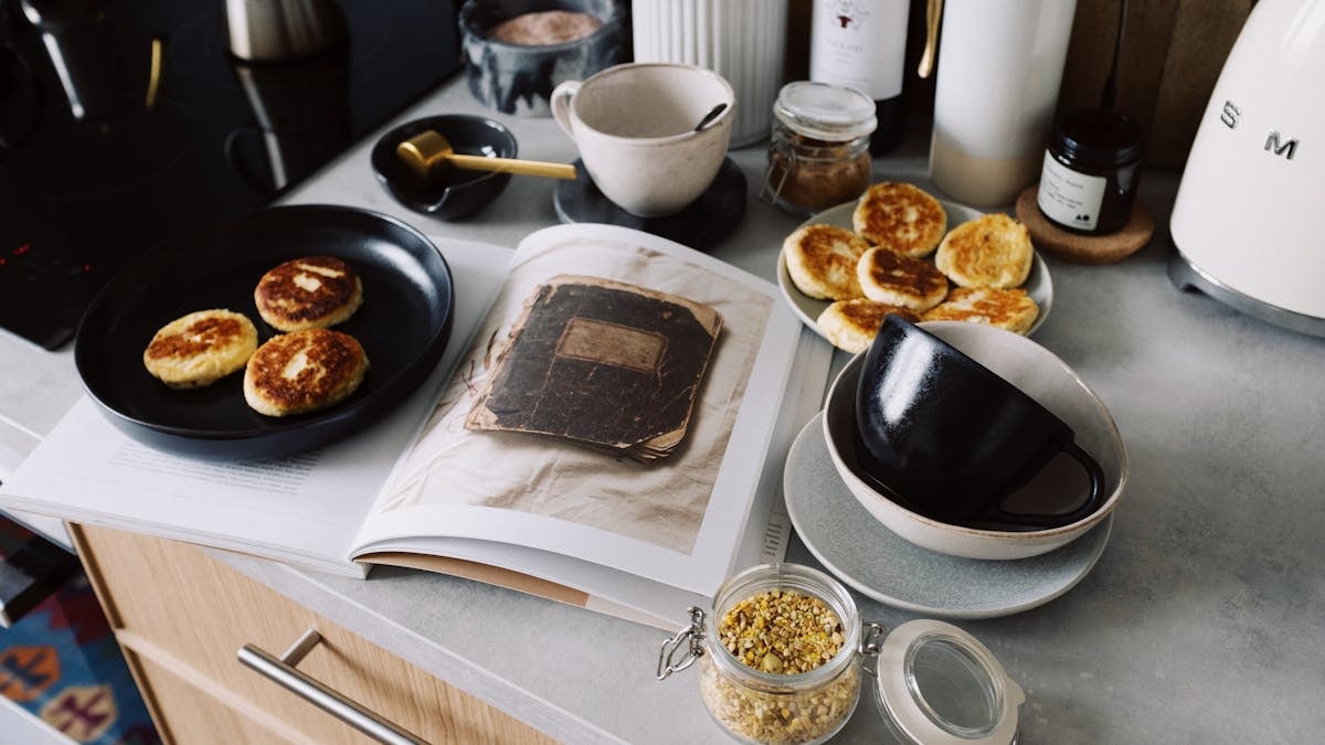 High angle of cookbook and cooked fritters on plates placed on table near utensils and ingredients in cozy kitchen at home